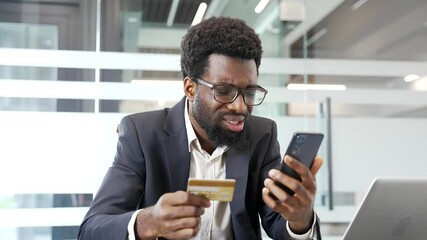 Shocked upset american businessman businessman checks his credit card on phone while sitting at workplace in business office. Frustrated black man faced fraud or scam. He became a victim of deception
