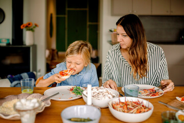 Boy eating pizza while sitting beside mother at dining table in home