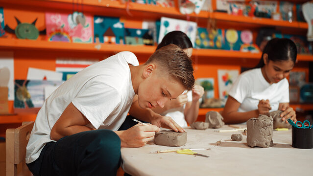 Highschool teenager using carving tool working at clay at pottery workshop. Group of happy diverse children working or modeling cup of clay with dough and equipment scatter around. Edification.