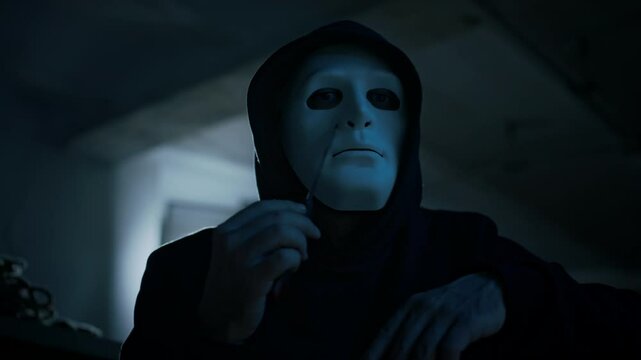 Lowkey closeup portrait of anonymous masked man in hood looking at camera while touching and checking sharp knife, passing it over face and neck