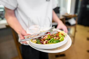 A waiter carrying a plate of fresh salad with mixed greens and garnishes. Glasses are visible in the background, suggesting a restaurant setting. Focus on the food presentation and service.