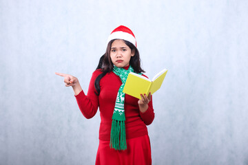 woman expression wearing red sweater red sweater with Santa hat and scarf celebrating merry christmas and new year, surprised to camera pointing right carrying book, isolated on white background