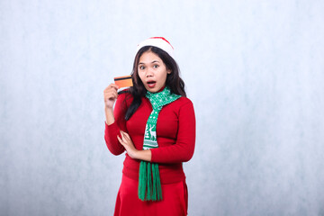 young asian woman shocked to camera holding credit debit card for payment, wearing red christmas sweater with santa hat and scarf, isolated on white background