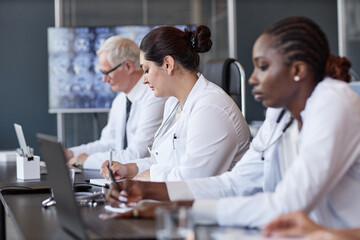 Obraz premium Side view of Middle Eastern female clinician listening to colleagues presentation keeping writing records while sitting at meeting table in medical office, copy space
