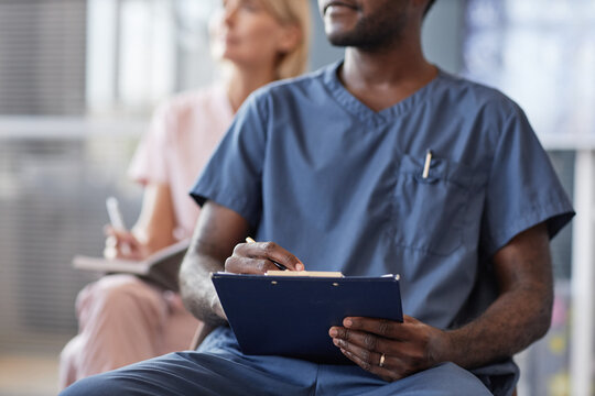 Cropped shot of African American man in blue scrubs keeping records of meeting on clipboard attending lecture in conference room while taking continuing learning course at hospital, copy space