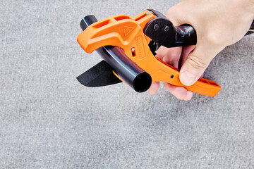 Hand of plumber holds pipeline scissors cutter with ratchet mechanism ready to cut segment of HDPE pipe, closeup on gray background.