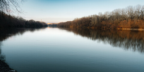 Tranquil river reflecting bare winter trees under a serene blue sky at sunset.