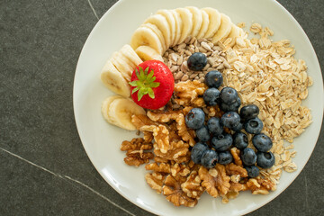 Healthy muesli ingredients on a big white plate, like oatmeal, walnut seeds, blueberries, banana slices, strawberry and sesame seeds, before adding yoghurt.
