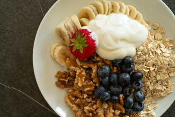 Healthy muesli on a big white plate, with oatmeal, walnut seeds, blueberries, banana slices, strawberry sesame seeds, and yoghurt.