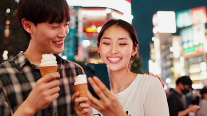 Couple, ice cream and happy with phone for travel, date or notification with bonding, love or contact in city. People, man and woman on smartphone, laugh or gelato on social network at night in Japan