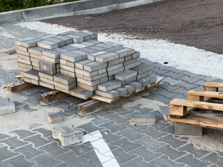 Sturdy concrete pavers are stacked on wooden pallets, surrounded by loose sand and gravel at an urban construction site near a paved street, ready for installation