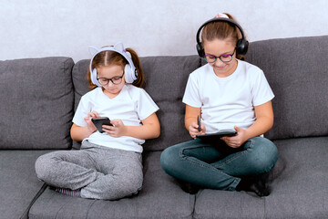 Two little sisters doing homework with a smartphone, sitting on the couch at home. A convenient and modern way to learn in a comfortable environment.