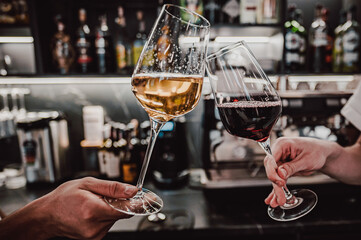 Two hands clink elegant wine glasses filled with sparkling white and red wine, set against a stylish bar background with various bottles, capturing a moment of celebration and joy.