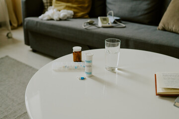 Various medication bottles, glass of water and open book arranged on white table in cozy living room with gray couch in the background