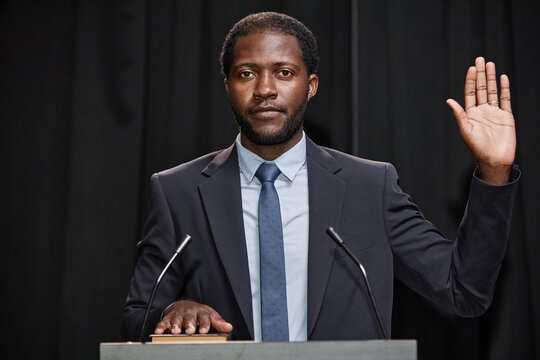 Portrait of African American male politician in suit with one hand on book and another raised swearing to uphold law while standing at podium on stage against backdrop of black curtains, copy space