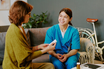 Smiling nurse wearing blue scrubs holding medical documentation while sitting on couch during...