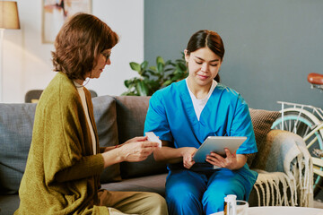 Elderly patient being assisted by caregiving professional sitting on couch in cozy living room setting without using natural light