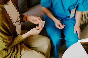 Assisting elderly patient with medication guidance, healthcare professional in blue scrubs providing support, senior holding medication blister pack with focused attention