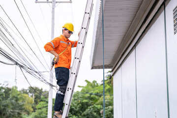 A safety conscious technician in an orange safety jacket and yellow hard hat ascends a metal ladder with tools clipped to his belt. The scene highlights outdoor work near a modern building facade. © ultramansk