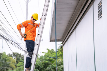 A safety conscious technician in an orange safety jacket and yellow hard hat ascends a metal ladder...