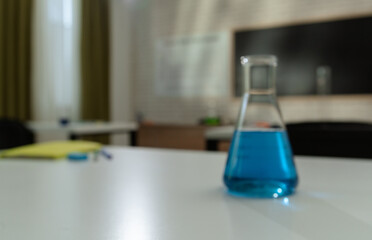 Empty school chemistry classroom with desks, chairs and chalkboard, workspace for teaching and studying. Glass bottle with blue liquid on table, close up.