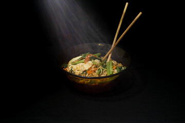 A bowl of noodles with mustard greens and egg, accompanied by chopsticks, is enhanced by dramatic lighting for a bold visual impact.
