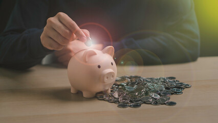 A hand places a coin into a piggy bank surrounded by coins, representing saving money, long-term investment, and financial planning for the future.