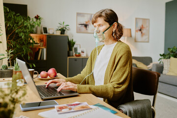 Elderly woman sitting at desk typing on laptop while wearing oxygen mask, potted plants and decor...