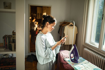 Young woman using smartphone while ironing clothes at home