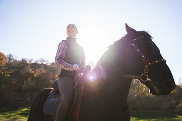 A young cheerful female equestrian riding a black horse in the countryside on a warm sunny autumn...