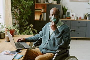 Elderly man sitting in wheelchair using oxygen mask while working on laptop in home office...