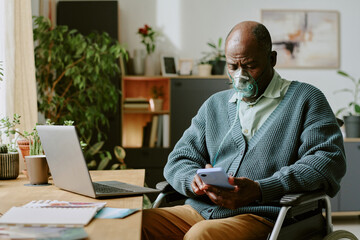 Senior man wearing oxygen mask while using smartphone and sitting in home office
