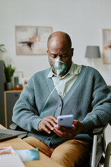 Portrait of elderly man sitting in wheelchair, wearing oxygen mask while using smartphone, looking...