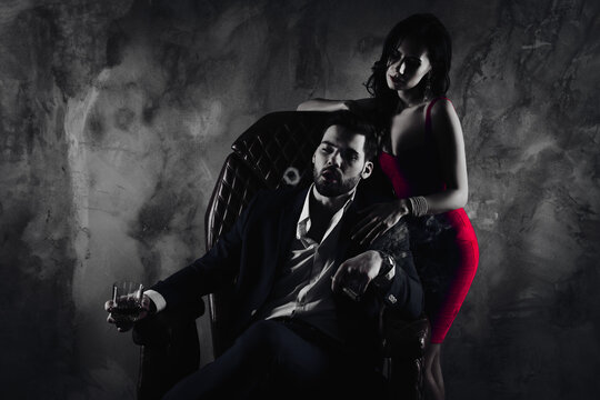 Portrait of virile boss in formalwear sitting in leather chair holding glass with whiskey cigar in hands making circles from fume charming lady standing near isolated on concrete background