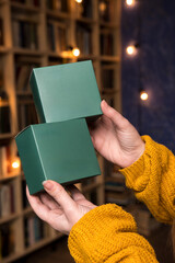 Close-up of hands in a yellow sweater holding two green boxes, with a blurred background of...