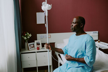 African American man sitting on hospital bed, holding IV drip and gazing out window, creating a...