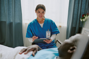 Nurse wearing blue scrubs is interacting with an elderly patient lying in a hospital bed, using a...
