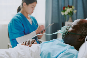 African American patient resting in hospital bed while being attended by Latin nurse writing...