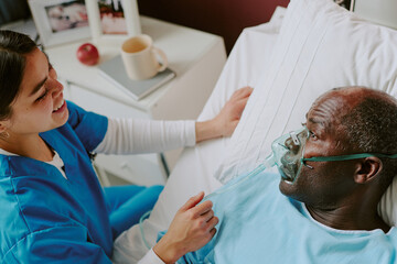 Nurse providing care for elderly patient lying in a hospital bed with oxygen mask, patient...