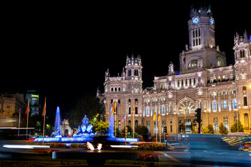 Fototapeta premium A large building with a fountain in front of it. The fountain is lit up at night. The building is surrounded by trees and cars