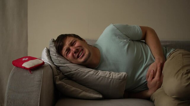 Man experiencing a painful stomach ache. He is lying on a sofa and holding his belly. First aid kit is visible on the sofa next to him
