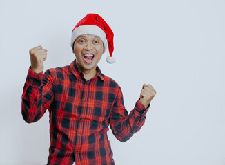 Merry young asian man wear a red checkered shirt and santa hat posing do winner gesture celebrate clenching fists say yes isolated on white background. New year 2025 holiday celebration concept.