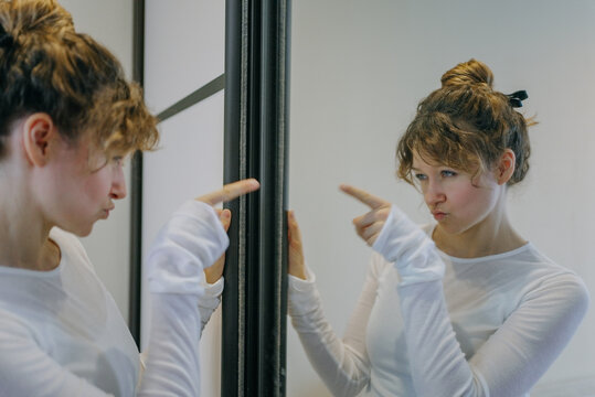 Young woman with curly hair tied in a bun, wearing a white long-sleeved shirt, points at her reflection in a mirror with a displeased expression