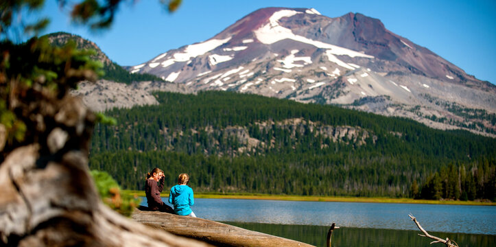 Sisters sitting on a log by a mountain lake with snow capped mountains in the background.