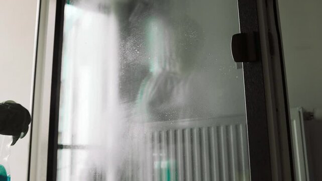 Person in rubber gloves sprays detergent and wipes window glass in room slow motion. Woman washes surface with cleaning supplies at home. High quality FullHD footage