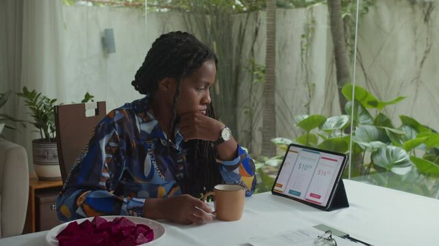 Black female manager looking at screen of digital tablet choosing between three subscription plans and drinking coffee while doing remote work