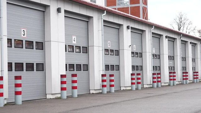 Valmiera, Latvia - November 28, 2024 - A row of gray industrial garage doors with red-striped bollards in front, part of a modern facility under a cloudy sky.
