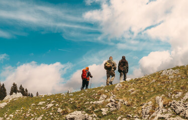 Three hikers with backpacks and outdoor gear walking up a grassy mountain slope under a vibrant blue sky, enjoying an adventurous trek in nature.