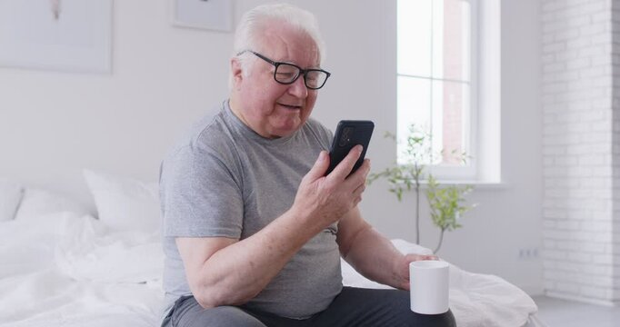 Positive elderly man sitting on a bed at home, drinking coffee and spending leisure time online using a mobile phone. Relaxation and entertainment through internet browsing in a bedroom.