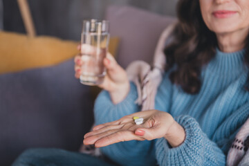 Elderly woman in sweater holding pills and glass of water, sitting on sofa in cozy home interior during daylight